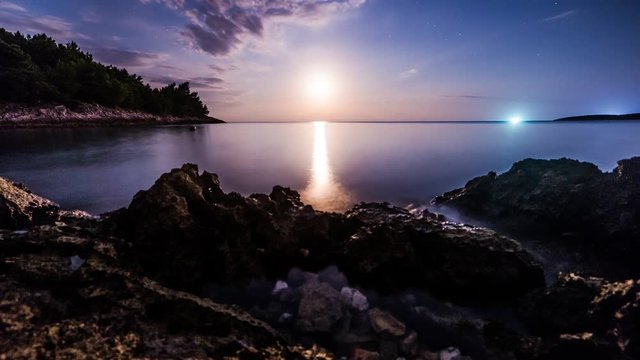 Time Lapse - Full moon / moonset at the  horizon of the ocean / sea with rocks in the foreground, the coast line and a bright light in the background