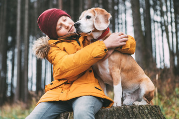 Two huging best friends portrait - boy and his beagle dog sit on the tree stump in the autumn pine forest.