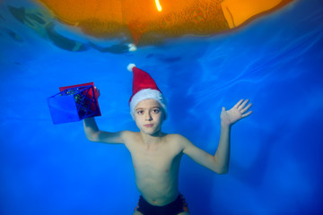 Obraz premium A little boy swims under the water in the pool in a red Christmas cap and with a gift in his hand on a blue background and looks at the camera. Portrait. Concept. Landscape orientation of the image
