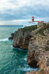 Fototapeta na wymiar Lighthouse on the top of the rocky cliffs with sea and clouds (Lighthouse of Cabo Sae Vicente, Lagos, Portugal, popular travel and holiday destination in Europe)