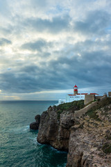 Fototapeta na wymiar Lighthouse on the top of the rocky cliffs with sea and clouds (Lighthouse of Cabo Sae Vicente, Lagos, Portugal, popular travel and holiday destination in Europe)