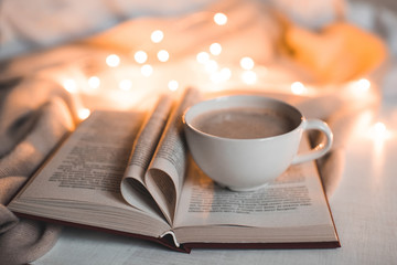 Cup of coffee staying on open book with heart shaped pages over lights at background. Cozy morning. Bedtime.