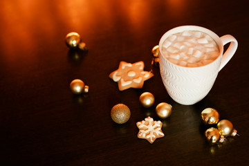 White cup of cocoa or coffee with hot chocolate and marshmallows and gingerbread cookies on the dark brown table and background with golden bokeh. Christmas holiday concept, free space for text