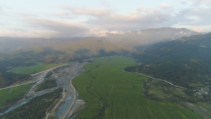Fototapeta premium Aerial view of farmer fields, river, village in the background of mountains at sunset on the island of Luzon, Philippines. Mountain landscape with valley and village.
