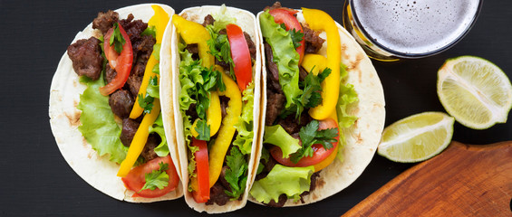 Tacos with meat and vegetables, beer and lime on a dark wooden background, overhead view. Flat lay, top view.