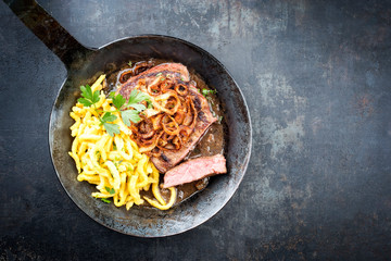 Traditional dry aged sliced roast beef with fried onion rings and Swabian spaetzle as top view in a wrough-iron pan with brown sauce with copy space