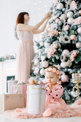 Merry Christmas and Happy Holidays! Cheerful mom and her cute daughter preparing for the Christmas. Mother decorates Christmas tree, having fun and playing together near Christmas tree indoors.
