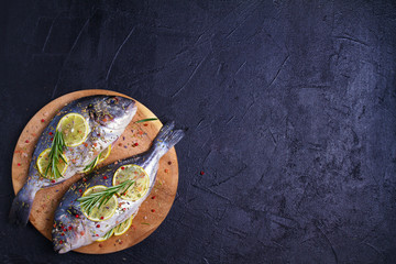 Obraz na płótnie Canvas Fresh uncooked dorado or sea bream fish with lemon, herbs, vegetables and spices on black background. View from above, top
