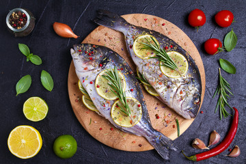 Fresh uncooked dorado or sea bream fish with lemon, herbs, vegetables and spices on black background. View from above, top