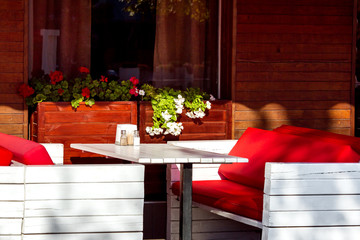 Summer terrace cafe with white wooden tables and red soft sofas to relax near the window with a flowerpot and blooms.