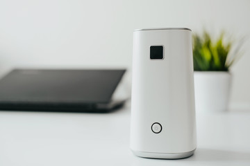 White air ionizer stands on the table, mlaptop and plant in the background. Concept of caring for fresh air in the apartment, in the office. Caring for well-being and health. Air filter.