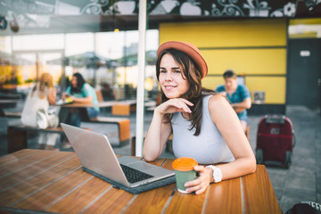 Fototapeta premium Beautiful young caucasian woman sitting on a terrace in a cafe in summer at a wooden table in a hat and a plate uses technology, working behind a laptop booking hotels trip in the summer
