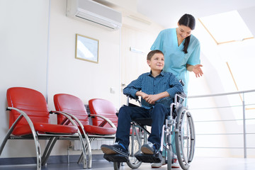 Young female doctor taking care of little boy in wheelchair indoors
