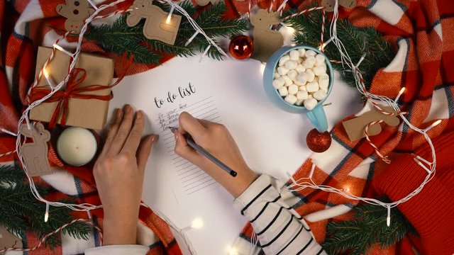 Woman hands writing new year to do list on white paper sheet, planning her future. Christmas decoration withfir branches, lights, red plaid. Flat lay. Top view. Mockup, copy space for your text.