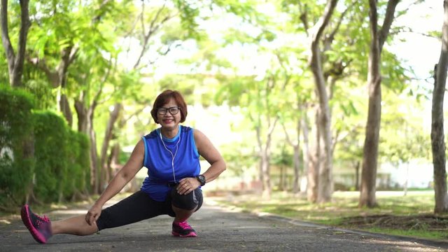 Senior asian woman stretch thigh muscles at park and listening to music. Athletic senior exercising together outdoor. Fit senior runners stretching before running outdoors.
