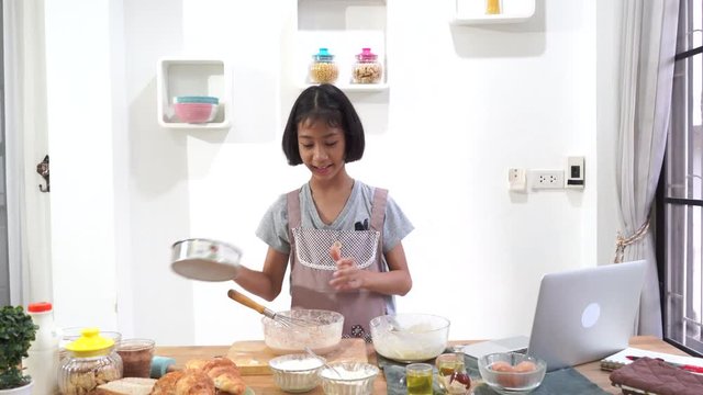 Asian little girl cooks learning online cooking using laptop computer in the kitchen at home