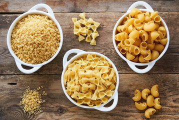 Various raw pasta in white bowls on a wooden background