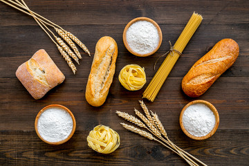 Homemade fresh bread and pasta near flour in bowl and wheat ears on dark wooden background top view