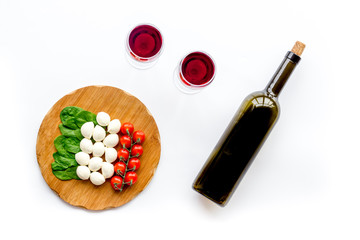 Italian cuisine, food concept. Italian flag made of mozzarella, tomatoes, basil on wooden cutting board near glass of red wine and wine bottle on white background top view