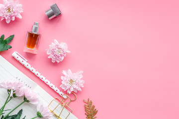 Perfume on feminine desk. Women's accessories. Perfume near notebook for dairy among flowers on pink background top view copy space