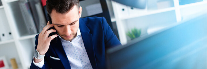 A young man is sitting in the office at the table, talking on the phone and holding a pencil in his...