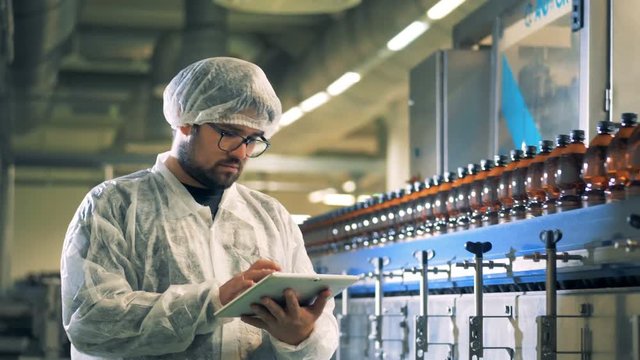 Brewery worker is operating a tablet while filled beer bottles are moving along the conveyor