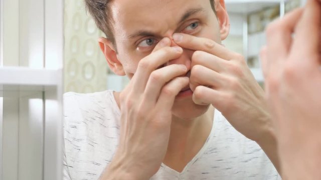 close up of young man standing close to a mirror in the bathroom and squeezing a pimple on his face.