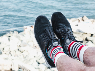Men's legs in sports shoes and white socks with a bright, sea pattern in the shape of anchors on the background of a rocky shore on a summer, sunny day. Concept of fashion, sport and recreation