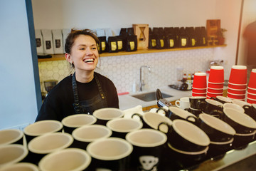 Local, small business concept - portrait of cheerful laughing female barista waiting for client while having job in cafe. Labor concept. View from above. A lot of empty cups in the foreground.