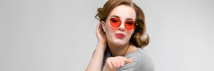 Charming young girl in a gray T-shirt on a gray background. Happy girl in red glasses. The girl is...