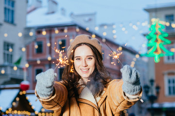 Amazing woman hat walking on the street with sparkler. Adorable female woman in winter outfit spending time outdoor and looking at Bengal light over Festive Christmas fair on background.