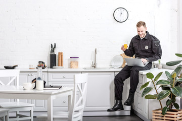 Young policeman sitting on table, drinking orange juice and using laptop at kitchen