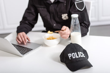 cropped view of police officer using laptop and eating cornflakes at kitchen table