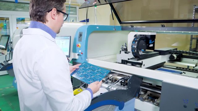 The engineer in a white robe and glasses inspect ready motherboard. PCB Processing on CNC machine, Production of electronic components at high-tech factory