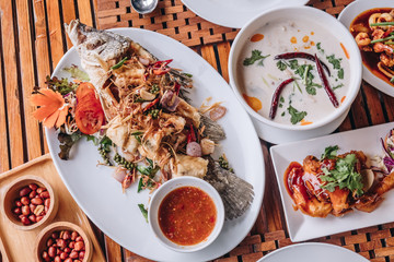 Thai dinner food with rice, Stir fried calamari and shrimp, Chicken soup with coconut milk, chicken grilled BBQ sauce, fried barramundi with herb on the white plate over wooden table in restaurant