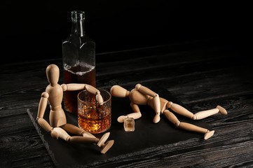Bottle, glass of whisky and two mannequins on wooden table against black background. Alcoholism...