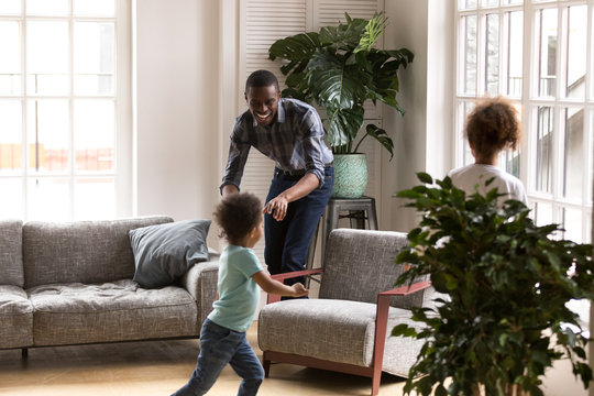 Happy African American family playing together at home, laughing father playing tag touch game with little preschooler daughter and toddler son, running in living room, catching to each other
