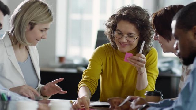 Cheerful Caucasian businesswomen writing down on sticky notes while having office meeting with multiethnic colleagues