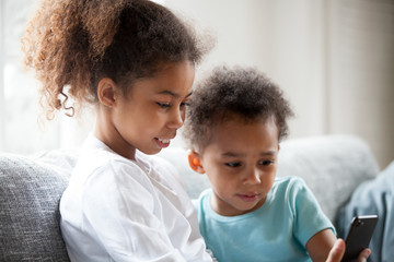 Pretty African American children using smartphone at home, little preschooler girl sitting together on couch with toddler boy, phone in hands, taking photo, making selfie, watching video close up