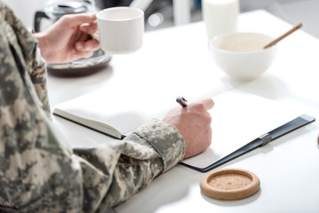 Fototapeta na wymiar cropped view of army soldier sitting at kitchen table, writing and having breakfast