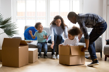 Happy playful large African American family moving in new apartment, little preschooler daughter sitting in cardboard boxes, father rolling her to mother, playing together, purchase property concept - obrazy, fototapety, plakaty