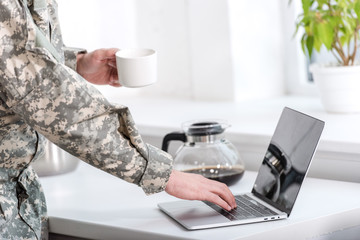 Fototapeta na wymiar partial view of soldier holding cup of coffee and using laptop at kitchen