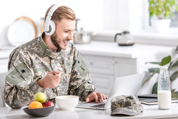 handsome soldier in headphones using laptop at kitchen table while having breakfast