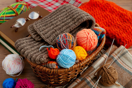 Yarn for knitting in the basket. Wicker basket with balls of yarn, knitting needles, blanket, book, glasses, bright shawl in a cozy home environment. Knitting as a kind of needlework.