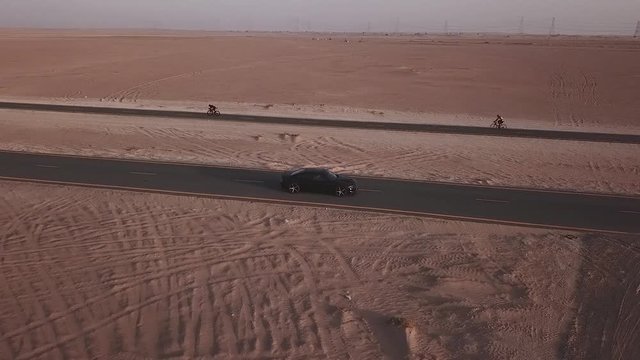 Car parked on road in the desrt with cyclist racing in the background in the desrt.