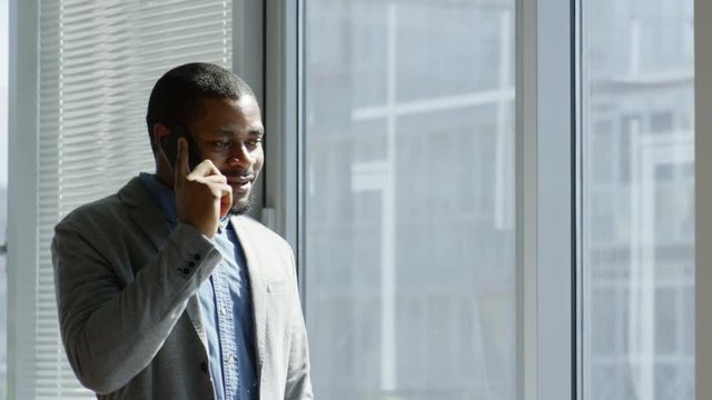 Tilt down of cheerful african american businessman standing by the window in office and chatting on mobile phone