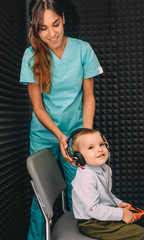 audiologist prepares little boy to hearing exam using headphones in special audio room.