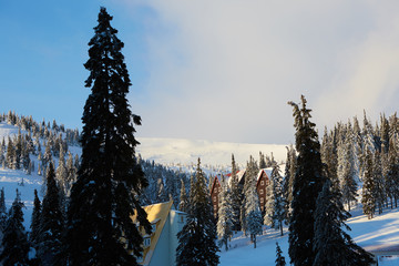 Mountain ski resort winter landscape photo. Tall fir trees and chalet houses covered with snow. Vacation and holiday cottages on sunrise in valley near ski elevator. Hotel buildings in the morning.