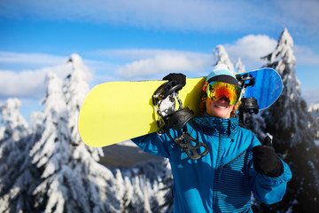 Smiling snowboarder posing carrying snowboard on shoulders at ski resort near forest before...