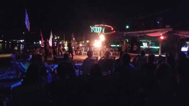 A guy fire twirling on a beach bar at night on Phi Phi island Thailand.
Audience watching as he tosses the burning stick in the air and catches it.